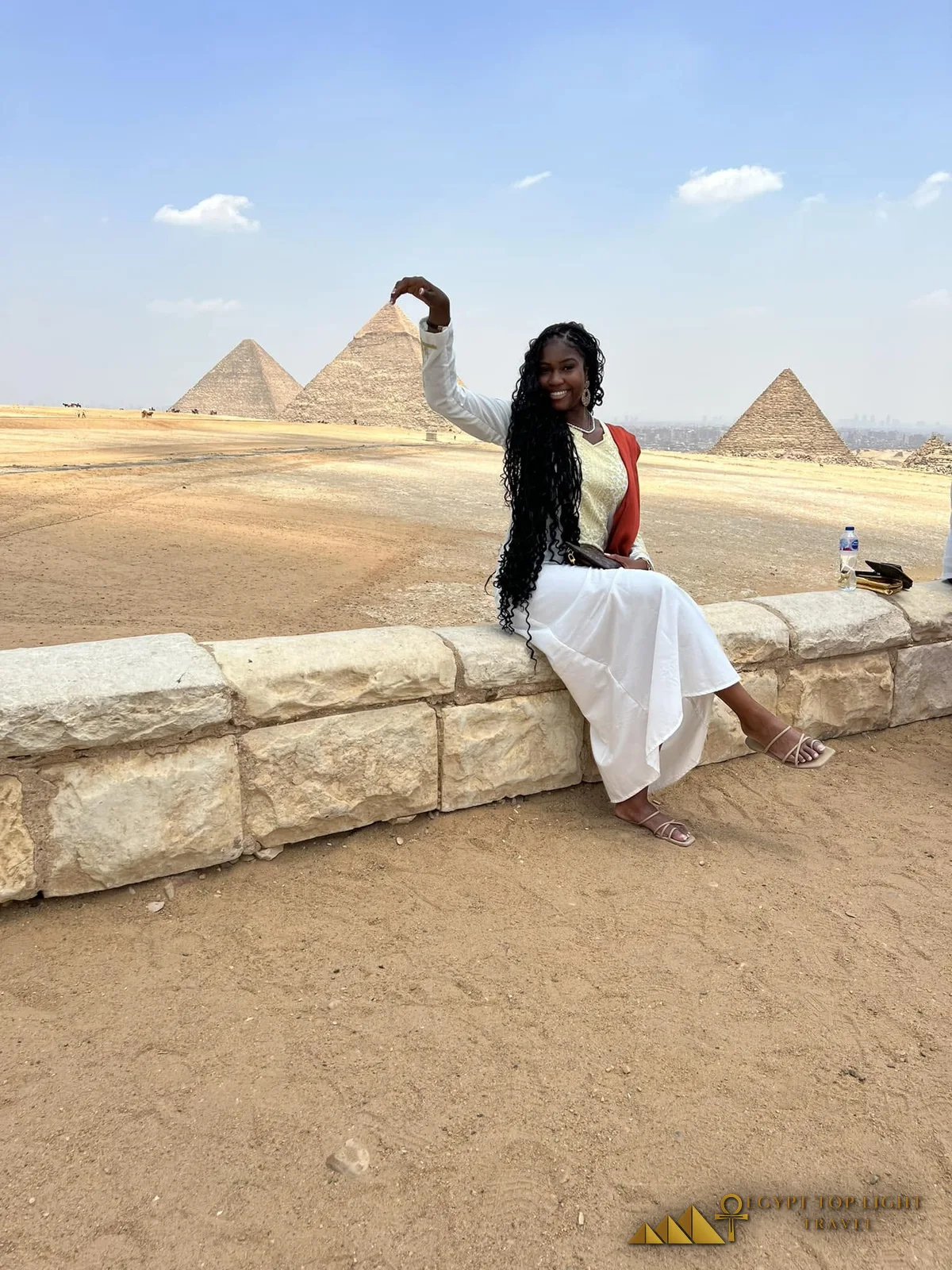 Black woman in a white dress posing playfully with the Pyramids of Giza